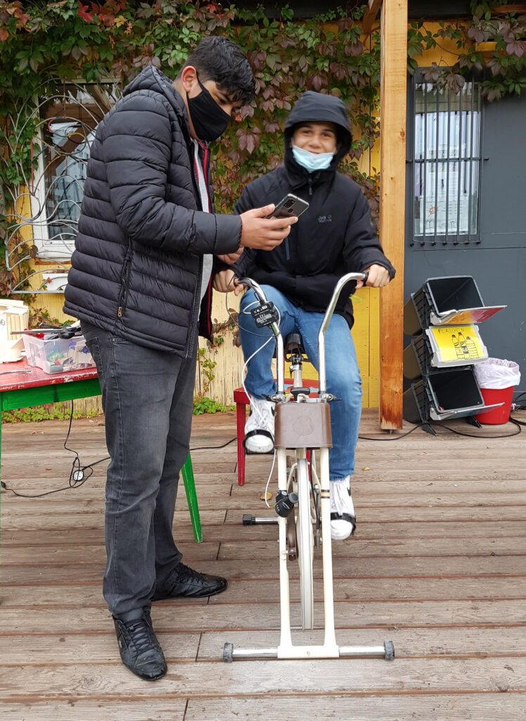 zwei Burschen sind zu sehen. Einer sitzt am Hometrainer der zu einer Handyladesation umgebaut wurde und tritt in die Pedale. der andere steht daneben und betrachtet sein Smartphone.